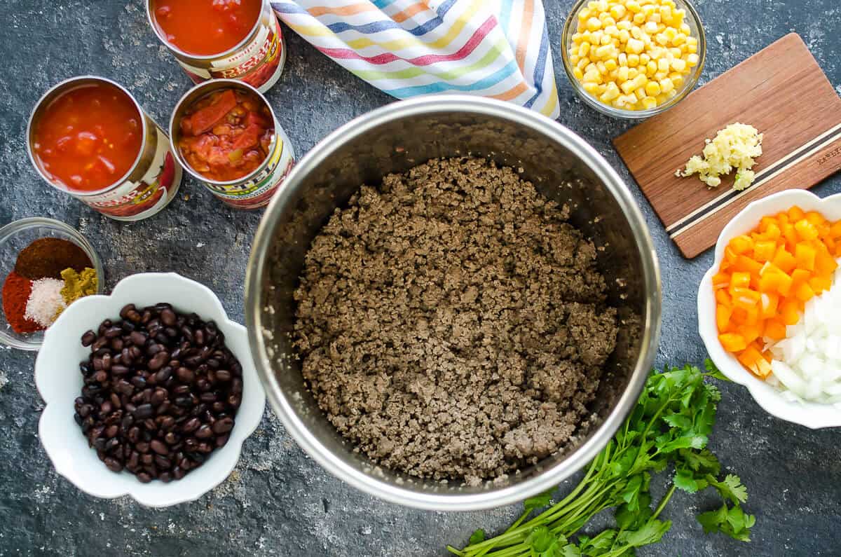 insert of instant pot filled with ground meat, other taco soup ingredients surround it, black beans, corn, diced tomatoes, and spices