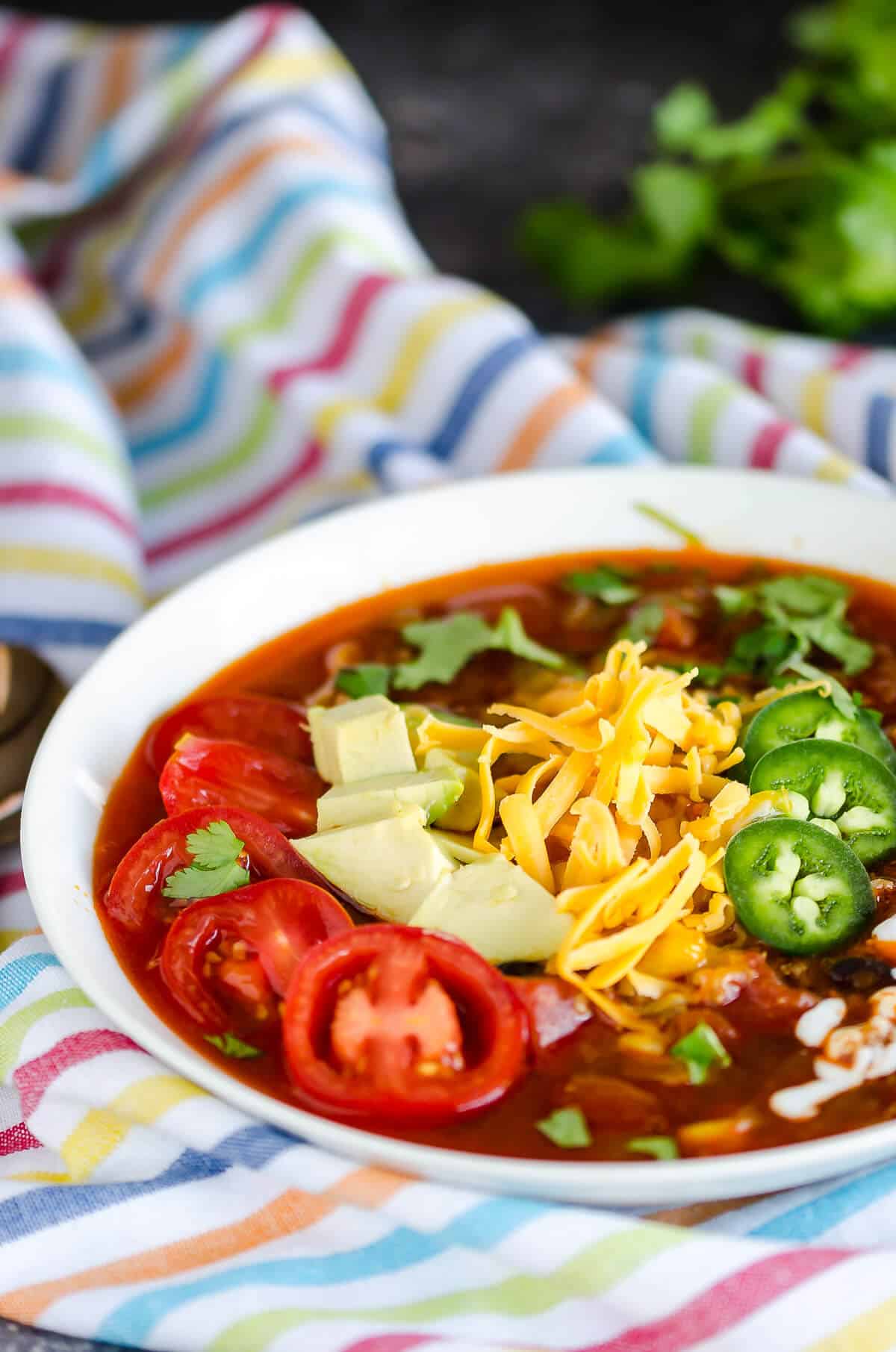 White bowl brimming with slow cooker taco soup, garnished with tomatoes, avocados, shredded cheese and jalapenos.