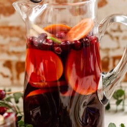 Clear glass pitcher filled with non alcoholic red sangria mocktail, fresh grapefruit, oranges, persimmon, and cranberries