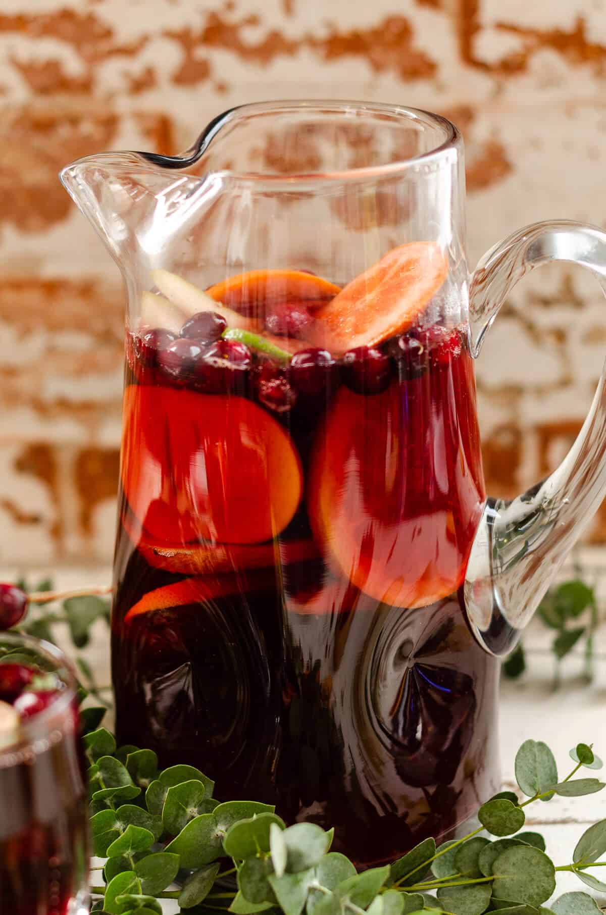 Clear glass pitcher filled with non alcoholic red sangria mocktail, fresh grapefruit, oranges, persimmon, and cranberries
