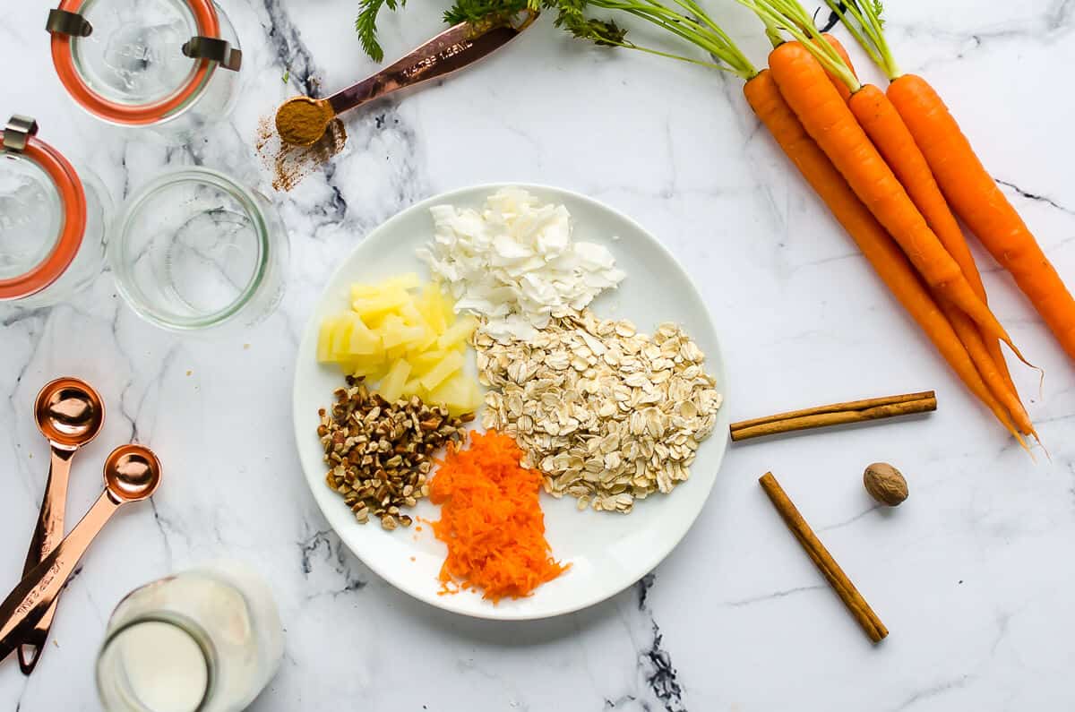 rolled oats, coconut flakes, chopped pineapple,pecans and finely grated carrots arranged on a white plate.