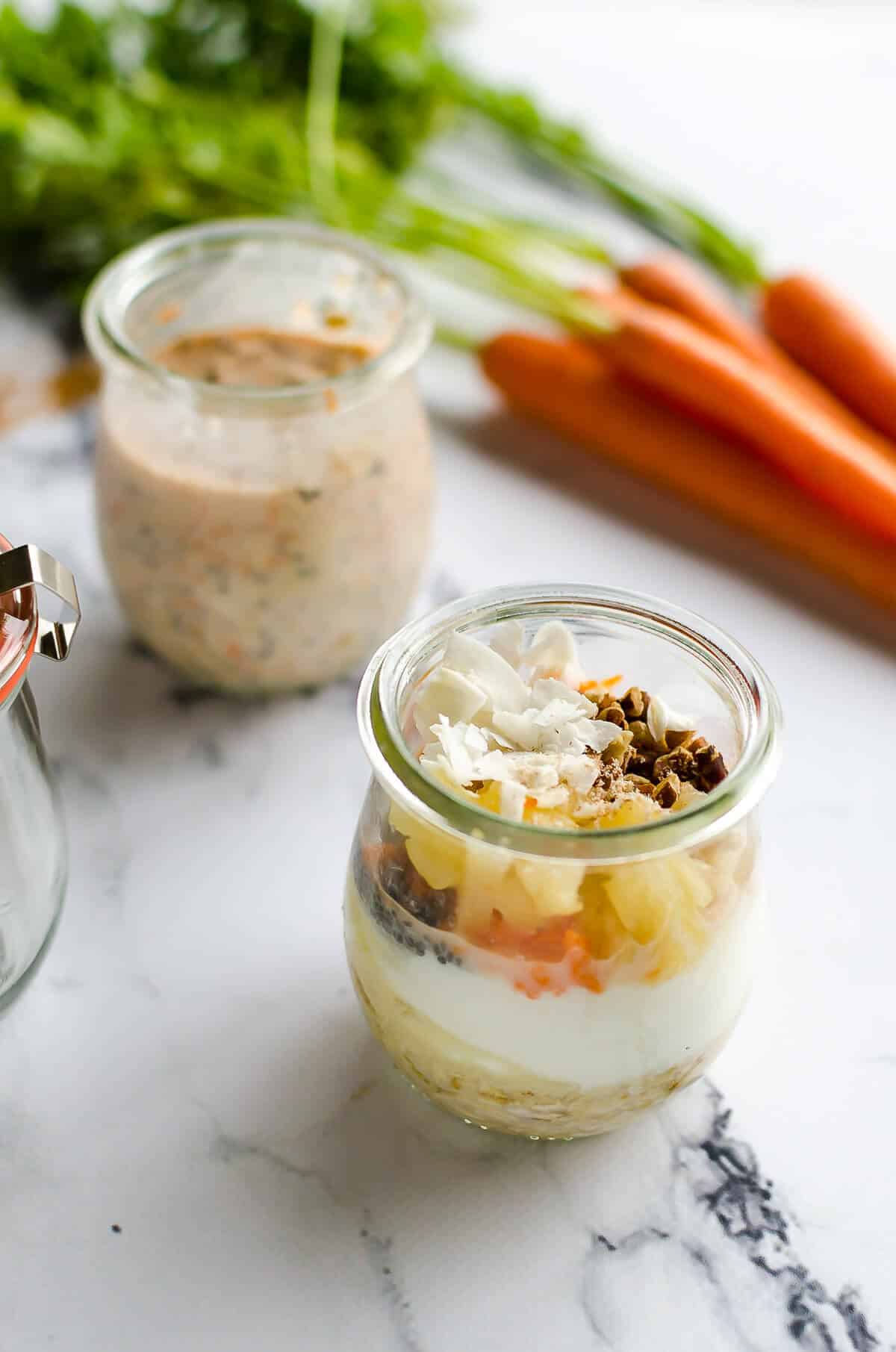 2 jars filled with carrot cake overnight oats ingredients, one stirred, one not stirred, includes yogurt, milk, oats, pineapple, coconut, carrots and pecans