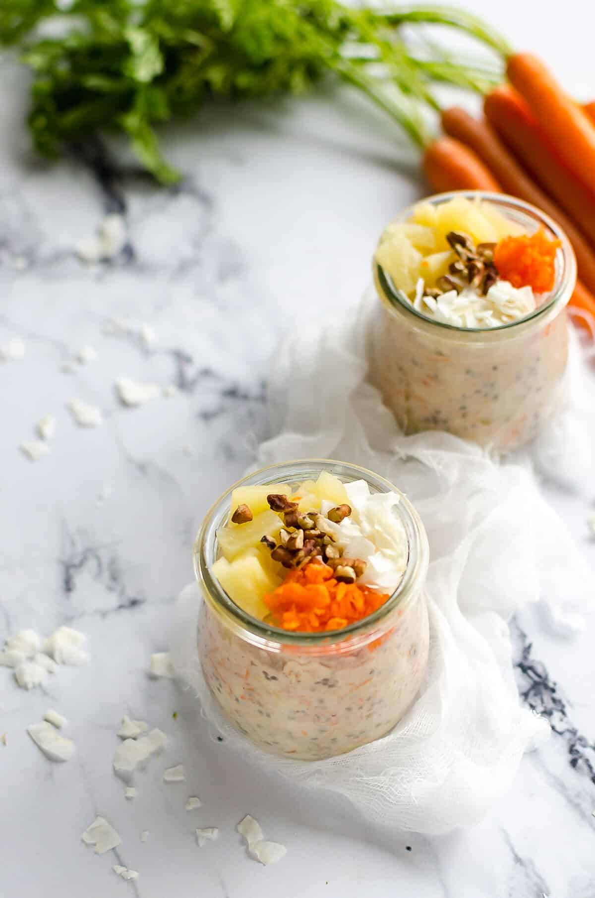 two jars full of carrot cake overnight oats, topped with pecans, pineapple, coconut and grated carrots