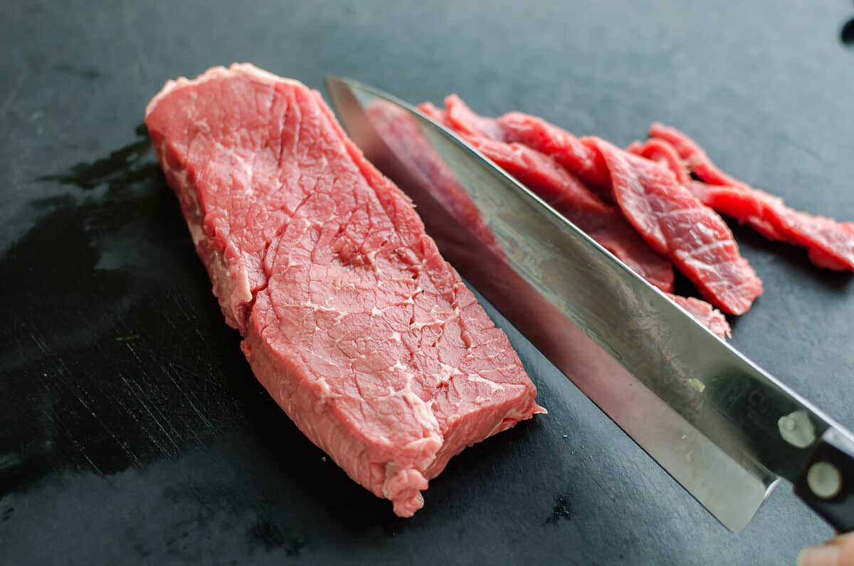 raw top loin steak being sliced into thin slices for philly cheesesteak