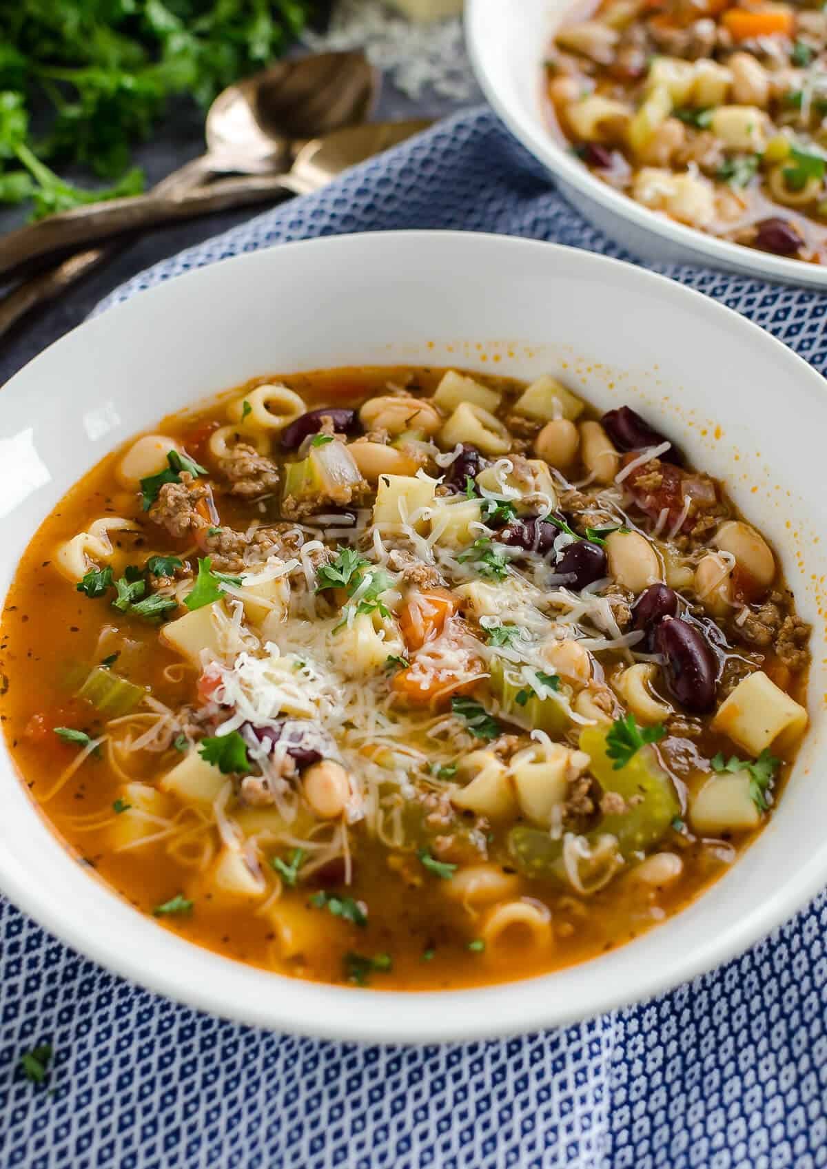 white bowl of soup with ground beef, kidney beans, carrots, celery and short pasta garnished with parmesan cheese.