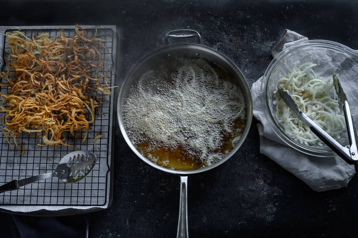 golden crispy onions on a rack next to pan of frying onions next to bowl of raw dredged onions