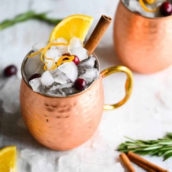 non-alcoholic cranberry moscow mule in a copper mug, garnished with orange peel, orange wedge, and cinnamon stick