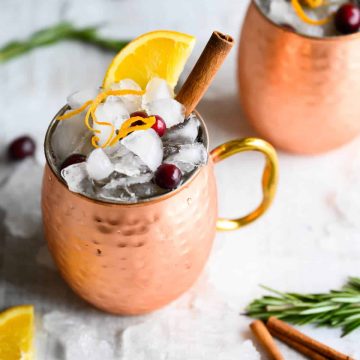non-alcoholic cranberry moscow mule in a copper mug, garnished with orange peel, orange wedge, and cinnamon stick