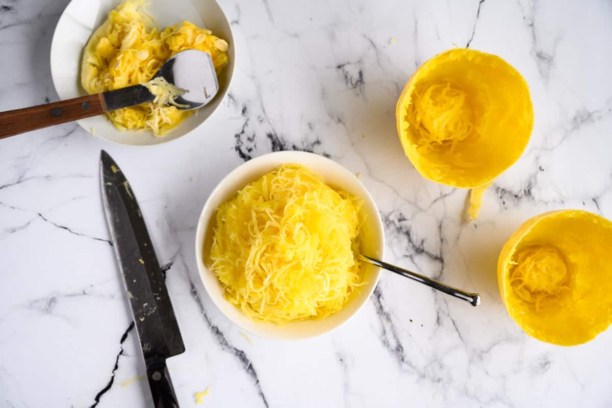 bowl full of cooked spaghetti squash noodles along side the empty skins and a bowl of the seeds.