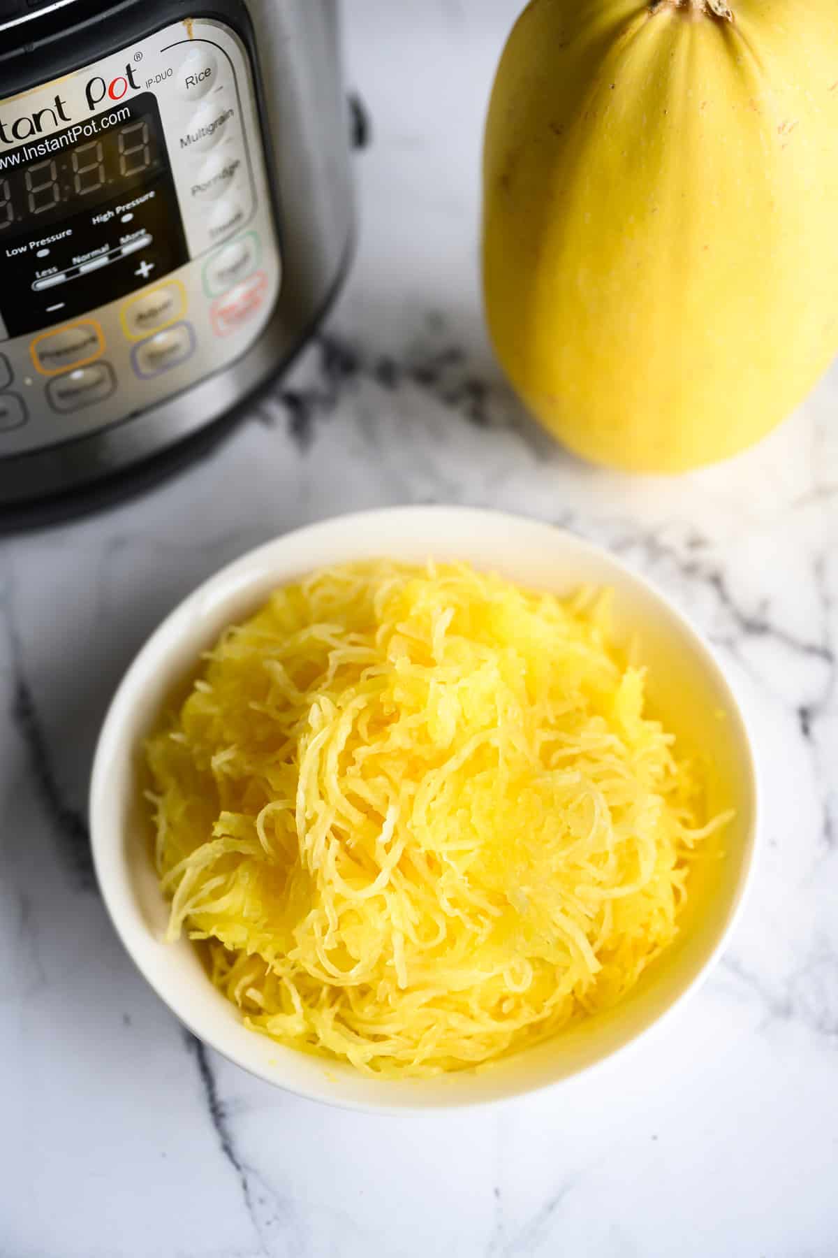 Instant pot next to a whole spaghetti squash and a bowl full of cooked spaghetti squash noodles