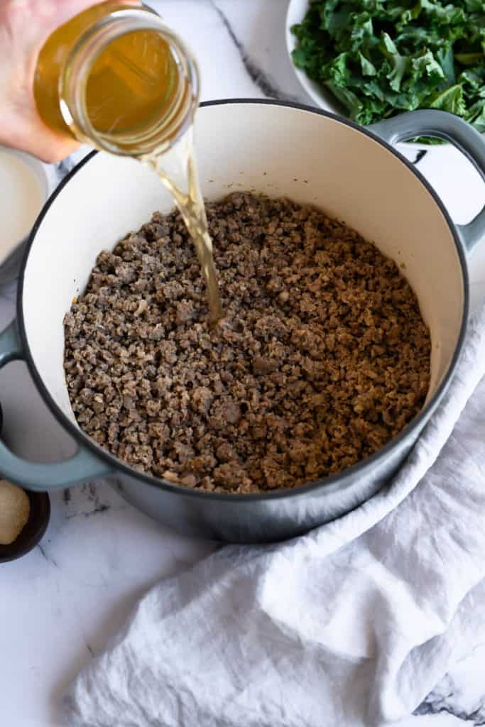 chicken broth being poured into a dutch oven full of cooked ground sausage