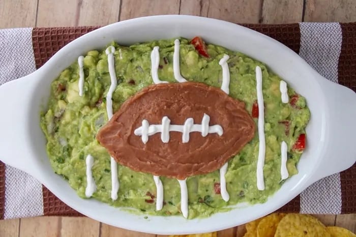 oval dish filled with guacamole, bean dip on top in the shape of a football, stripes of sour cream on the guacamole to look like a football field, and on the football to look like stiching.