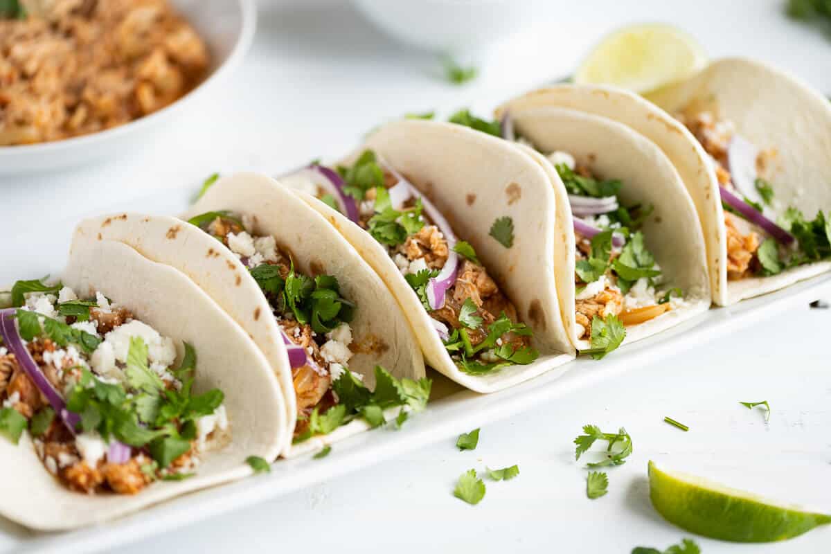 chicken tinga tacos on a platter garnished with red onion, cotija cheese, cilantro and lime