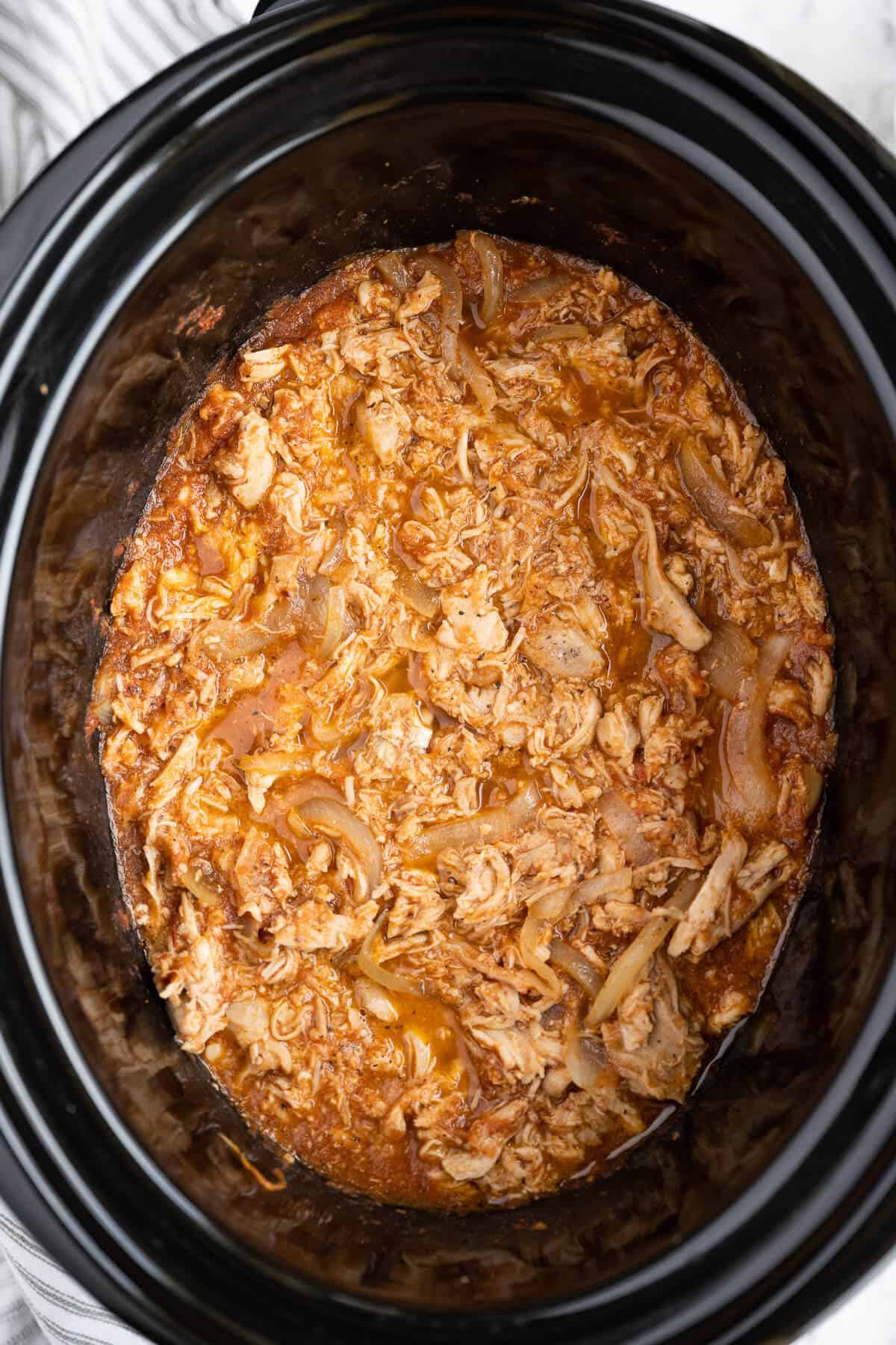 shredded chicken and sliced onions simmering in a tomato chipotle pepper sauce in a crockpot