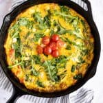 cooked frittata in a cast iron pan garnished with arugula, rosemary, and diced tomatoes