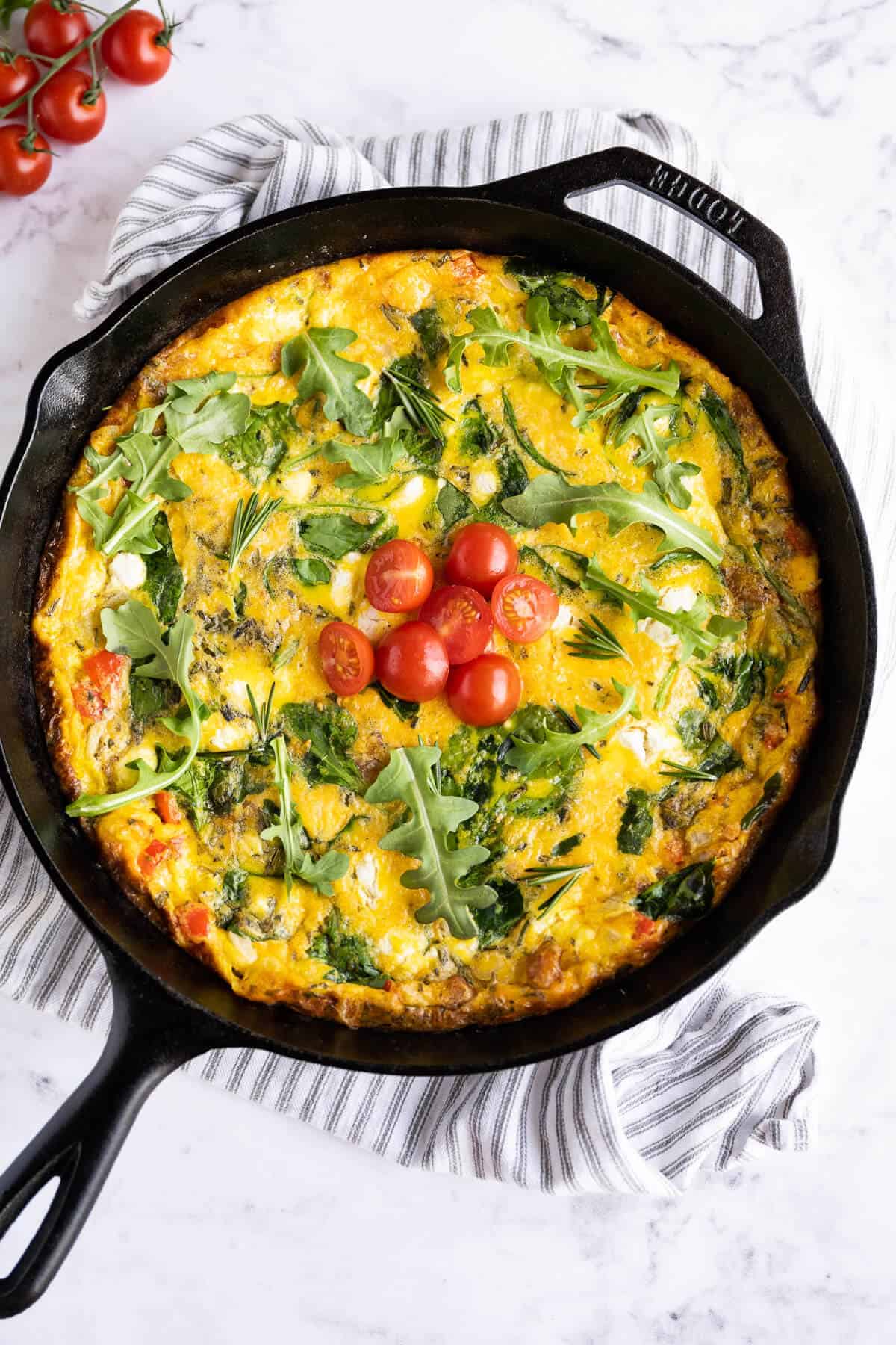 cooked frittata in a cast iron pan garnished with arugula, rosemary, and diced tomatoes