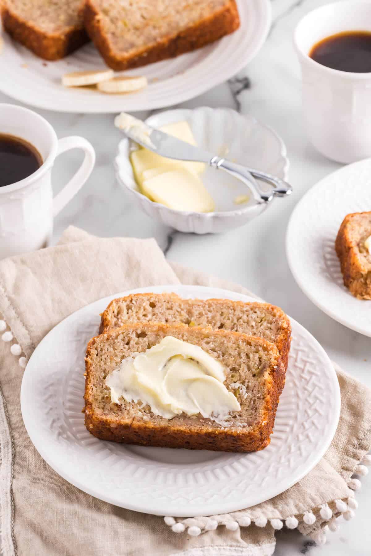 Two slices of banana bread on a plate surrounded by other plates of banana bread, a dish of butter, and cup of coffee.