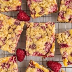 Squares of raspberry crumble bars on a cooling rack. A few fresh raspberries in between the bars.