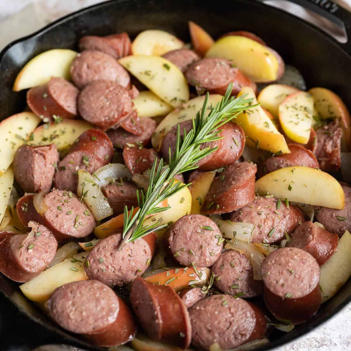 Sliced kielbasa, apples, and onions in a cast iron skillet with a rosemary spring on top.