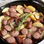 Sliced kielbasa, apples, and onions in a cast iron skillet with a rosemary spring on top.