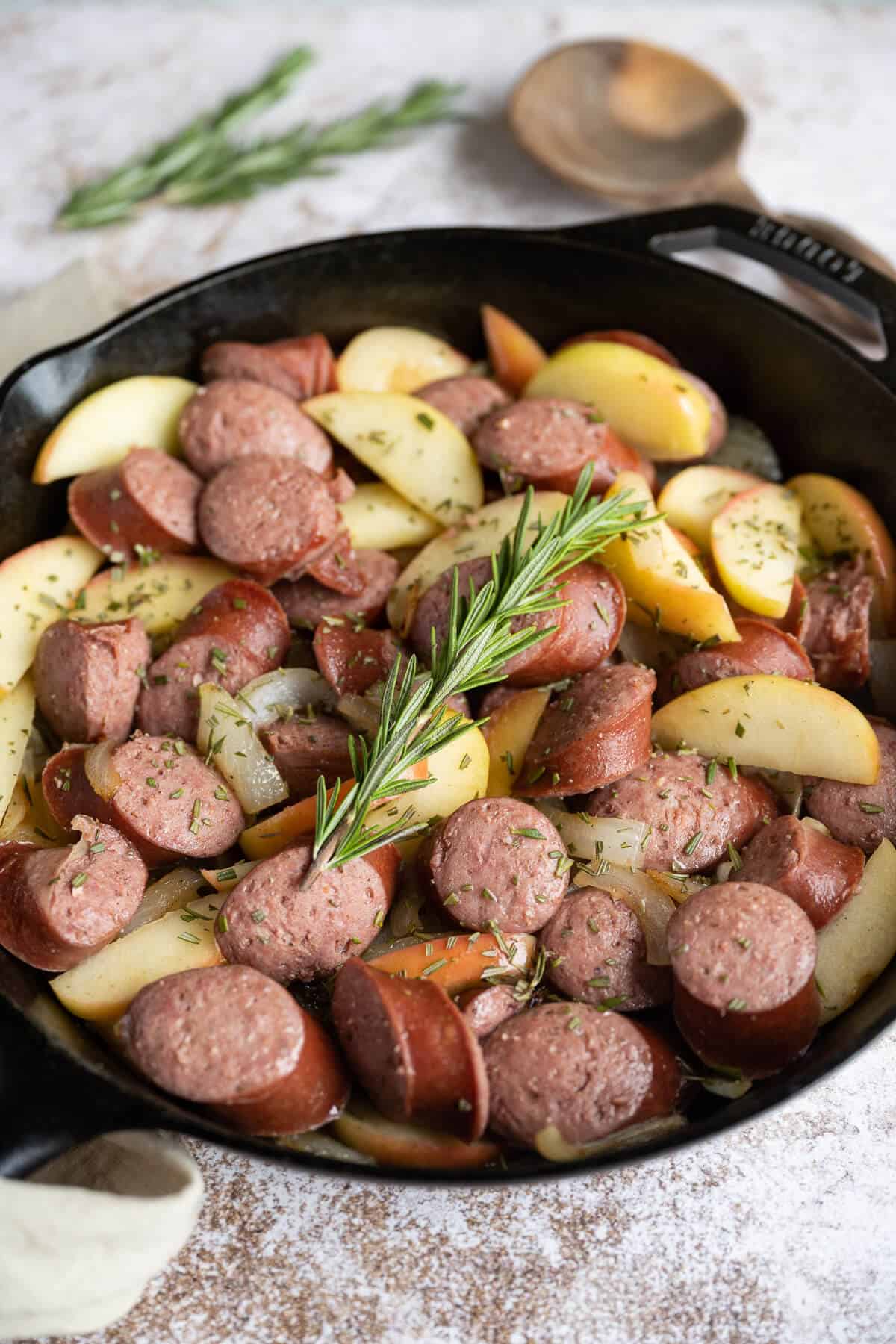 Sliced kielbasa, apples, and onions in a cast iron skillet with a rosemary spring on top.