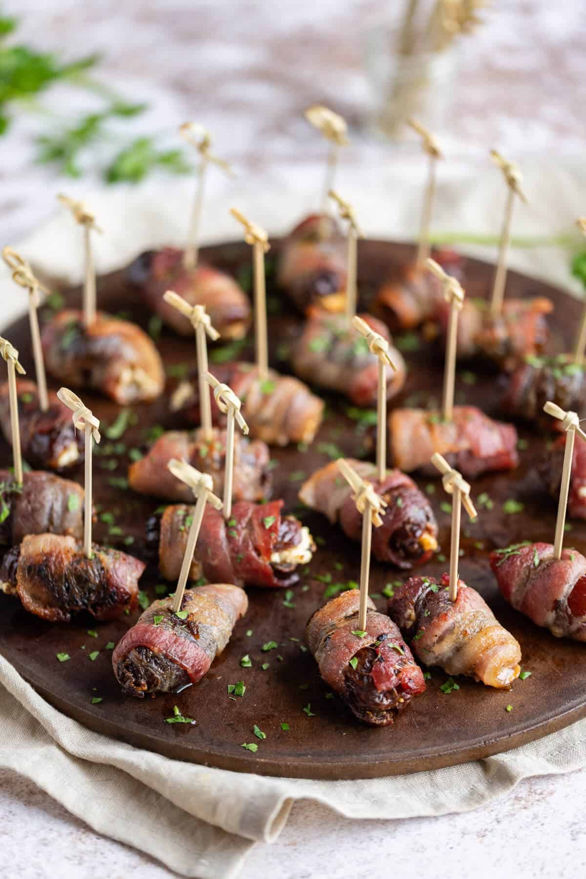 Bacon Wrapped Dates poked with cocktail skewers on a round serving tray garnished with fresh chopped parsley.