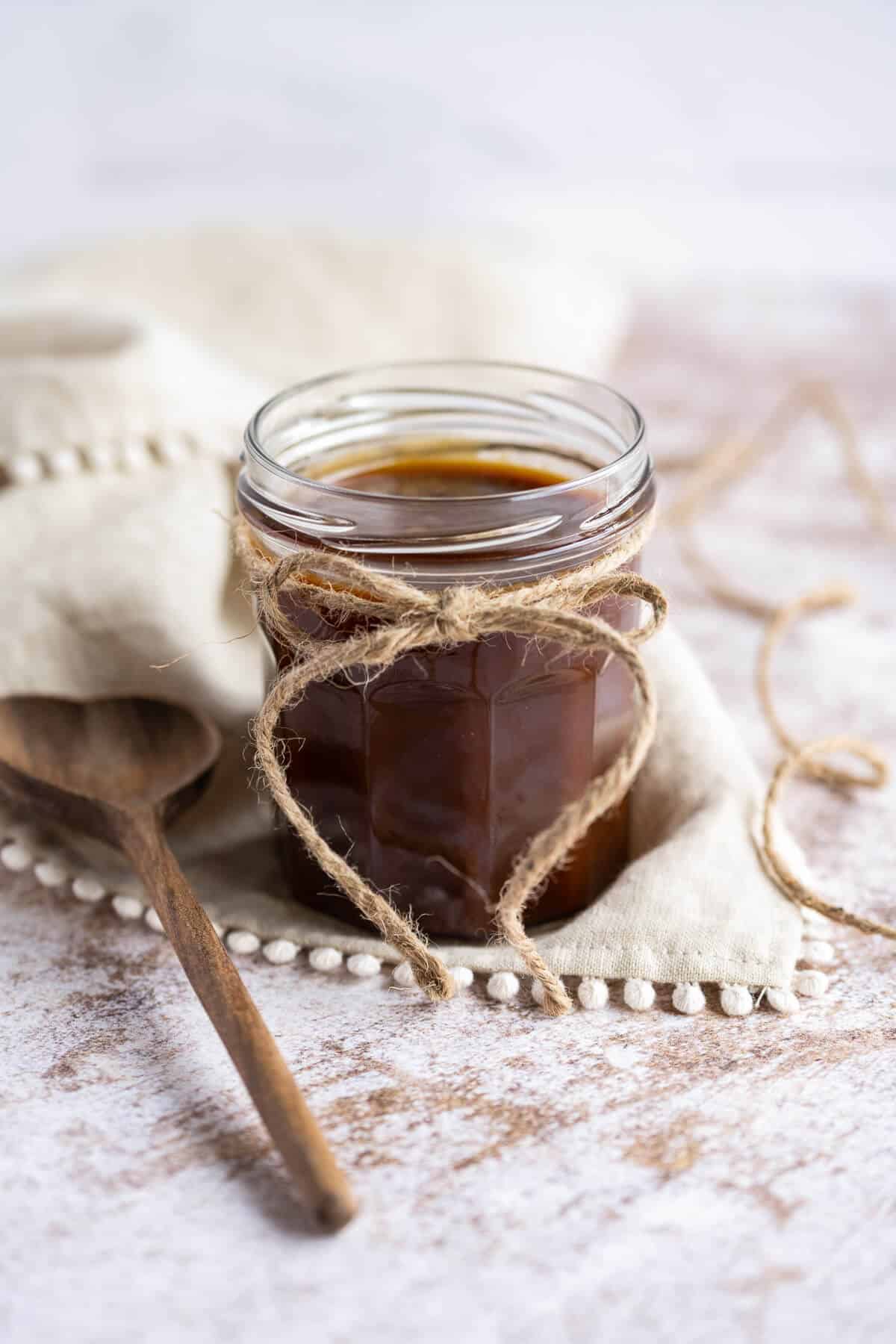 Homemade caramel sauce in a clear glass jar with a ribbon around it. A wooden serving spoon next to it.