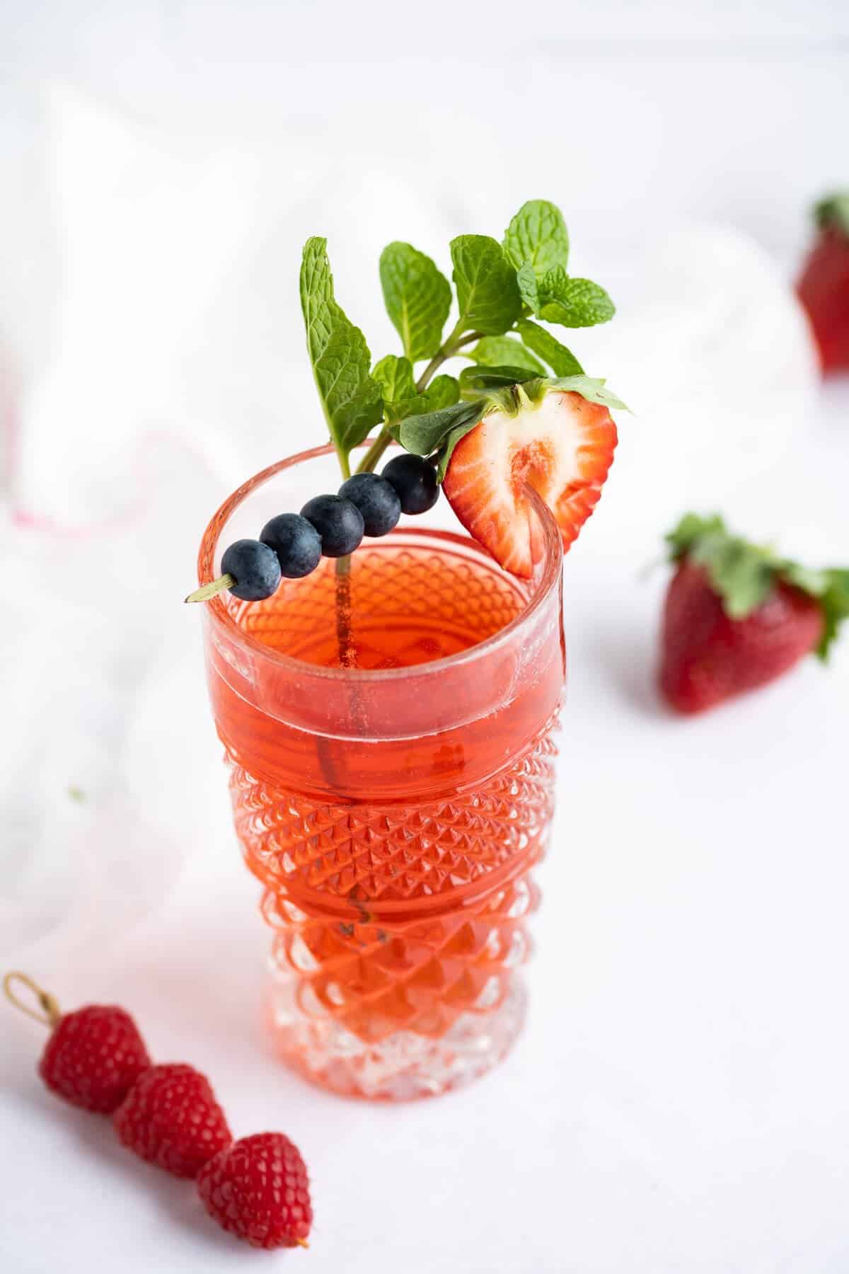 Berry flavored kombucha in a decorative tall glass garnished with a cocktail skewer of blueberries, a sliced strawberry, and a sprig of mint leaves.