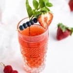 Berry flavored kombucha in a decorative tall glass garnished with a cocktail skewer of blueberries, a sliced strawberry, and a sprig of mint leaves.