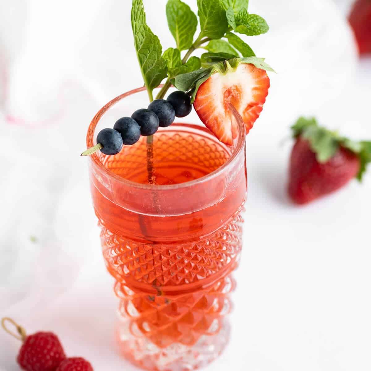 Berry flavored kombucha in a decorative tall glass garnished with a cocktail skewer of blueberries, a sliced strawberry, and a sprig of mint leaves.