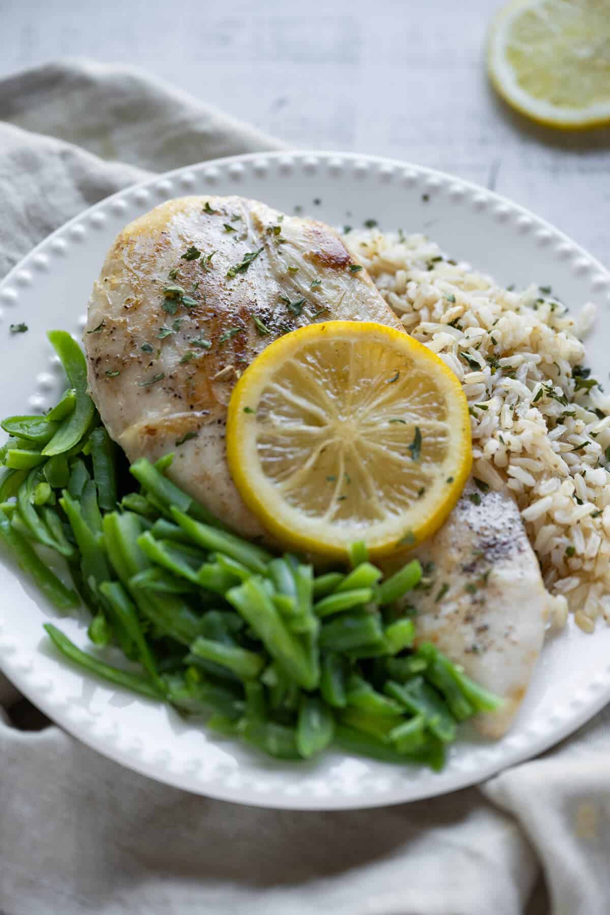 Cooked boneless chicken breast garnished with a lemon slice on a bed of rice with green beans on the side.