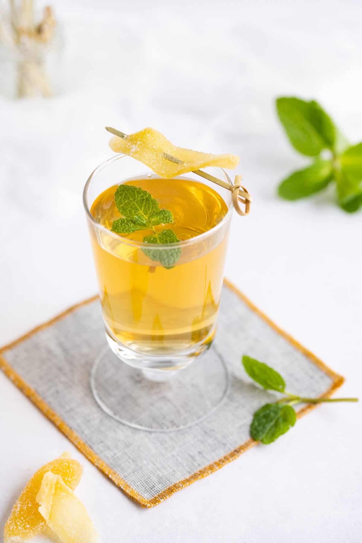 Glass of ginger kombucha garnished with a mint leaf and crystallized ginger on a cocktail pick.