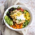 Mexican grilled chicken on top of quinoa and veggies, garnished with cilantro, cheese and sour cream in a white bowl.