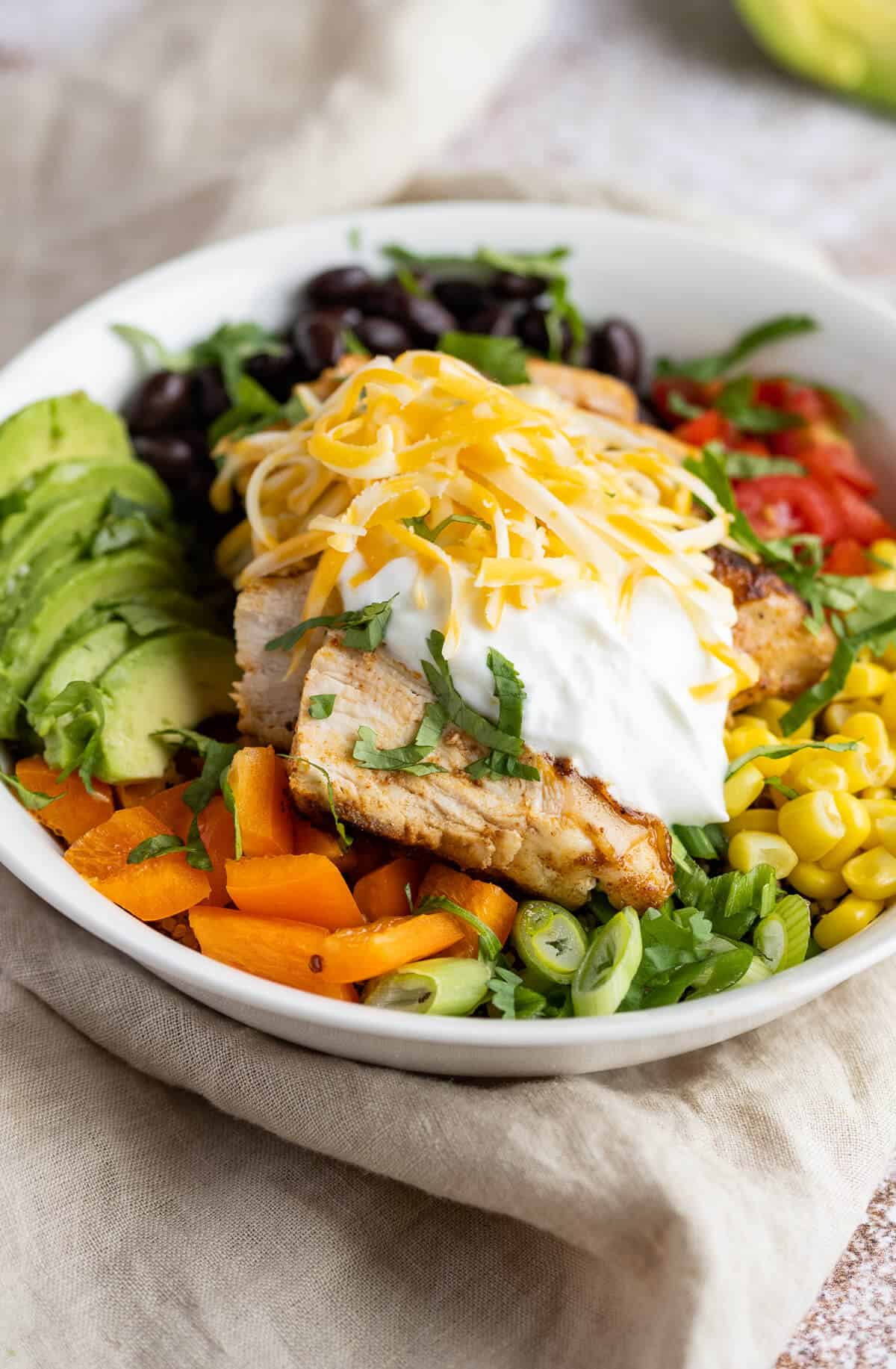 Bowl of Southwest grilled chicken on top of a bed of quinoa, black beans, corn, tomatoes, peppers, and avocado, garnished with cheese and sour cream.