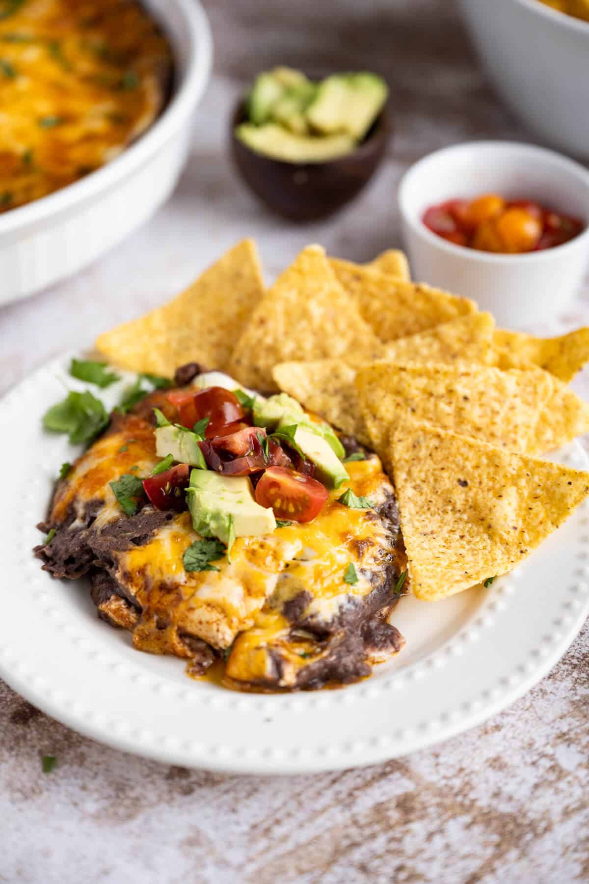 Warm black bean dip topped with melted cheese, cilantro, diced tomato and diced avocado, on a white plate with a side of tortilla chips.
