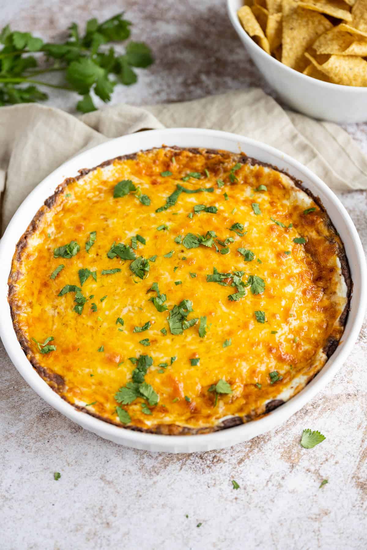 Warm black bean dip smothered in melted bubbling cheese in a round pie plate. Cilantro is sprinkled over the top and it is next to a bowl of tortilla chips.