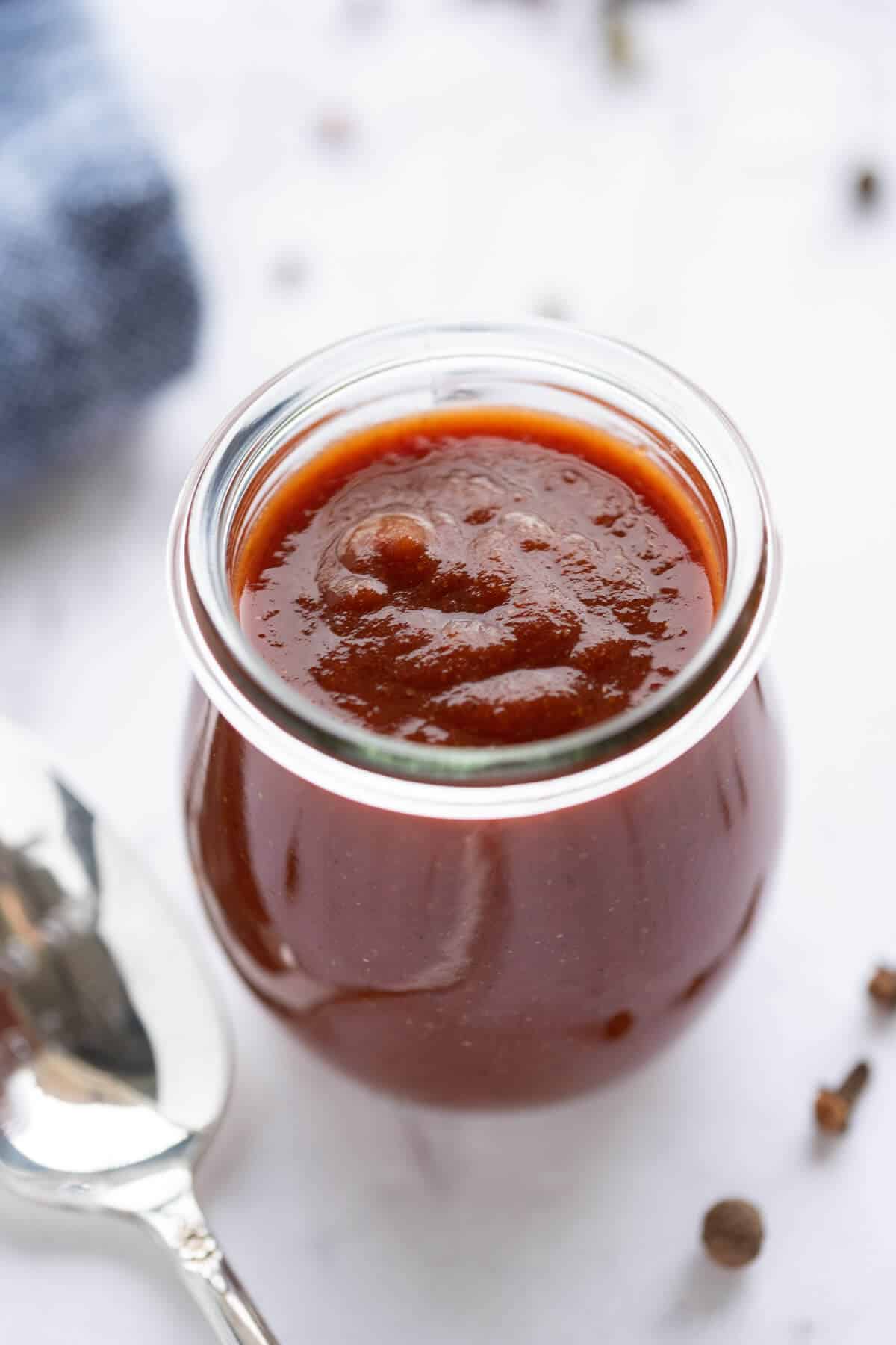 Sugar free barbecue sauce in a glass Weck jar with a spoon beside it. Blue napkin in the background, allspice and peppercorns in the foreground.