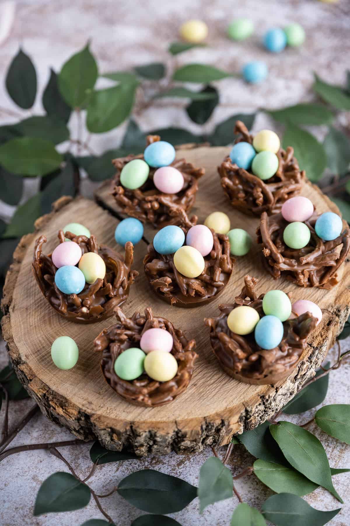 A group of chocolate birds nests filled with mini candy coated eggs on a wooden serving platter surrounded with greenery.