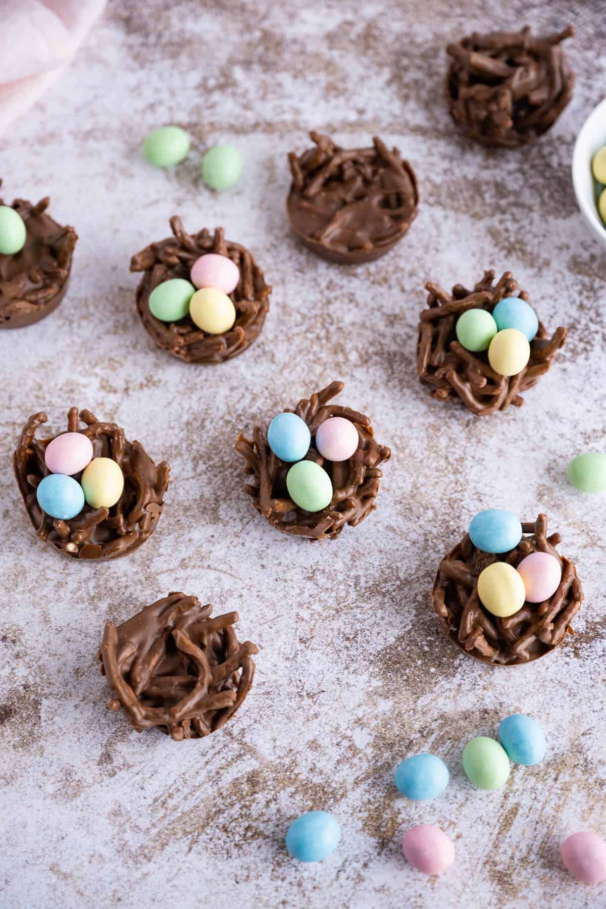 Several Chocolate Birds Nest Cookies, some filled with candy eggs, some empty. Mini candy eggs scattered alongside them.