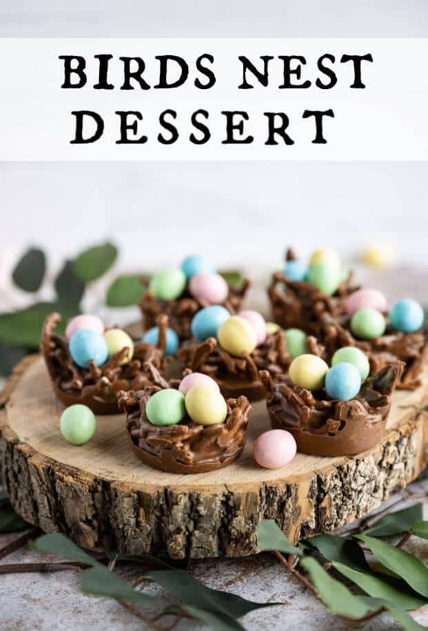 A group of chocolate birds nests filled with mini candy coated eggs on a wooden serving platter surrounded with greenery. via @artfrommytable