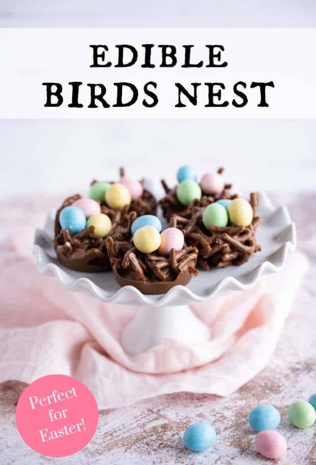 Several Bird Nest Cookies filled with pastel colored candy coated mini eggs on a pedestal. Greenery is surrounding it, a few candy eggs is in the foreground. via @artfrommytable