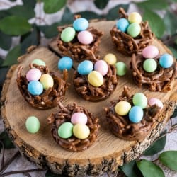 A group of chocolate bird nest cookies filled with mini candy coated eggs on a wooden serving platter surrounded with greenery.