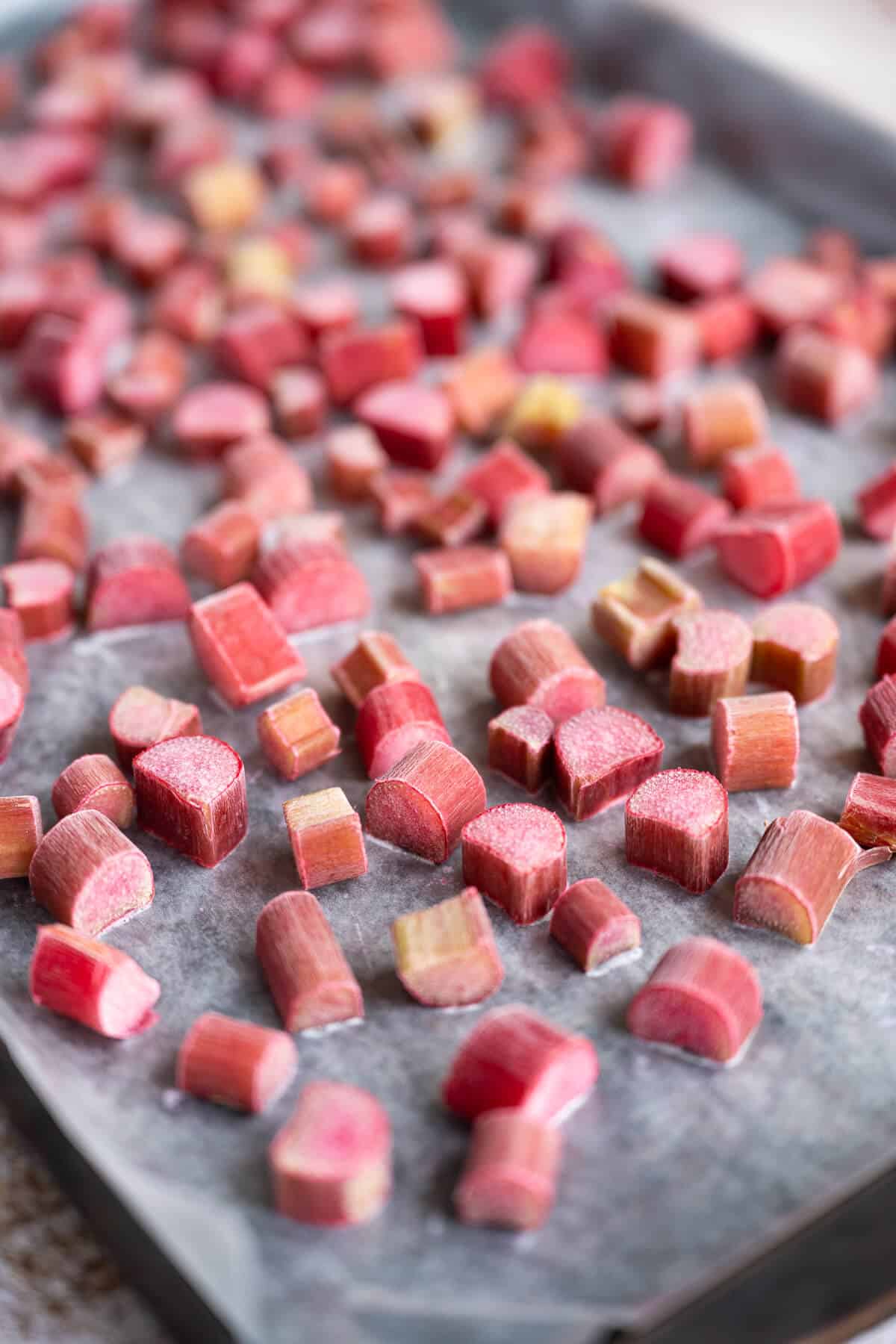 Frozen chunks of rhubarb on a wax paper lined rimmed baking sheet. Closer pieces are in focus, further pieces are out of focus.