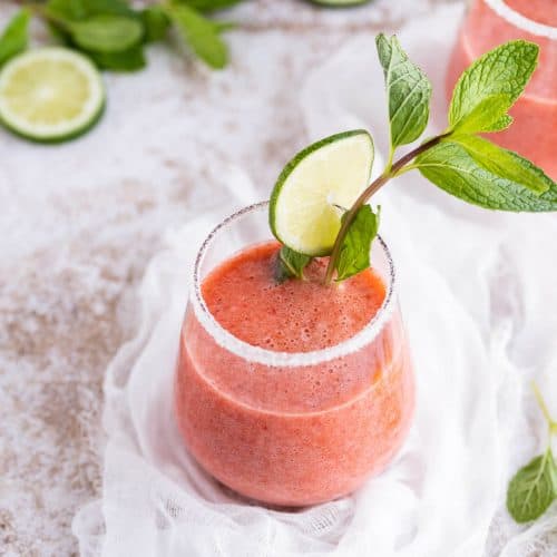 Clear sugar rimmed glass filled with a blended strawberry drink, garnished with fresh mint and a slice of lime.