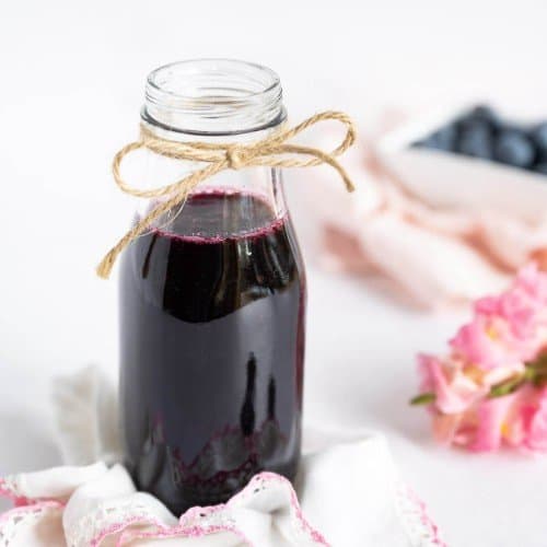 Blueberry simple syrup in a glass milk bottle type jar. A piece of natural twine is tied in a bow at the top of the jar. Blueberries and fresh flowers are in the background.