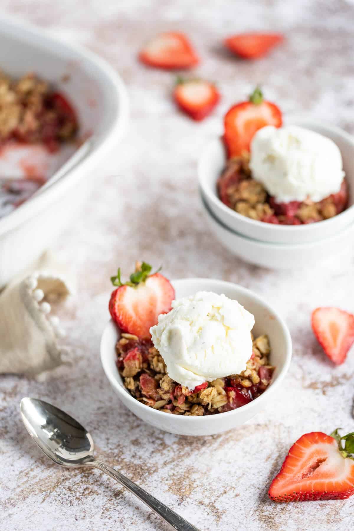 2 bowls of Rhubarb Crisp with strawberries topped with a scoop of ice cream. Each bowl is garnished with a fresh strawberry slice. A spoon is alongside one of the bowls.