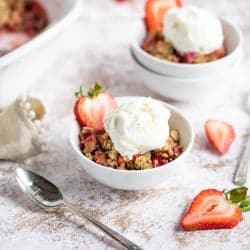 Two bowls of rhubarb strawberry crisp topped with ice cream and a fresh strawberry slice. A few slices of strawberries are scattered around.