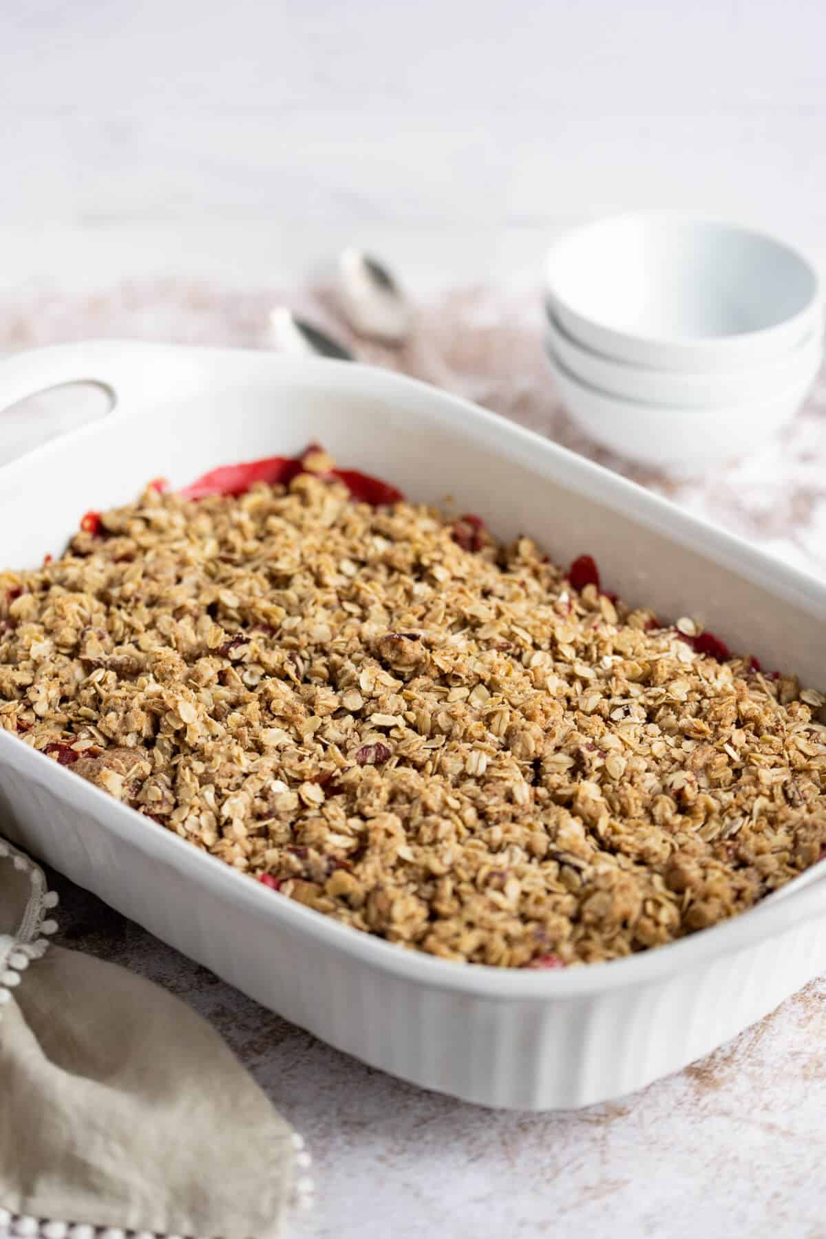 Rhubarb Crisp with Strawberries in a white casserole dish, just baked. Bowls and spoons are alongside it.