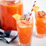 Glass of homemade strawberry lemonade with a red and white striped straw, and garnished with a strawberry slice and a lemon wedge. Behind the glass is another glass and a pitcher of the remaining lemonade.