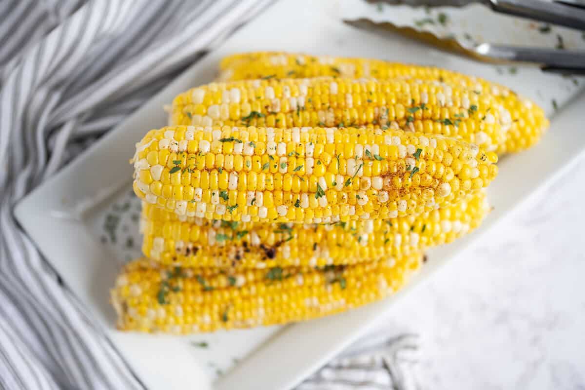 6 ears of grilled corn on the cob stacked on a white rectangle platter. A pair of tongs is in the background.